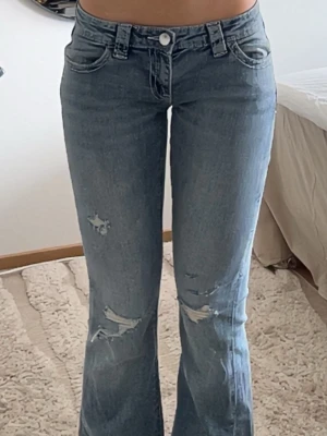 Blå bootcut jeans med slitningar - Säljer ett par ljusblå bootcut jeans med slitna detaljer på framsidan och snygga broderade fickor bak. Jeansen har låg midja och klassisk femficksdesign. Materialet är denim i bomull med lite stretch för skön passform. De är använda 1 gång!!