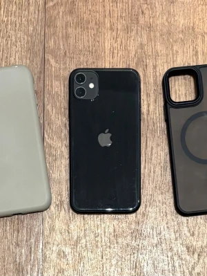 Svart iPhone 11 med två skal 64GB - Säljer en svart iPhone 11 med stilren glasbaksida och rundade kanter. Telefonen har dubbla kameror på baksidan och levereras med två olika skal – ett ljusgrått och ett svart med MagSafe-ring. Perfekt för dig som vill ha en snygg och pålitlig iPhone. 64GB. Snabb Leverans!