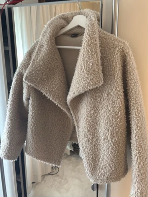 Beige teddyjacka med knappar  - Mysig beige teddyjacka. Jackan har en fluffig struktur och är tillverkad i ett mjukt, teddyliknande material. Perfekt för att hålla värmen och ge en trendig look. Passar till många olika stilar och är enkel att matcha, jackan är andvänd ett få antal gånger. Pris kan diskuteras 