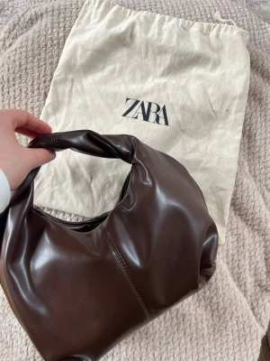 Mörkbrun elegant handväska från ZARA - Säljer en snygg mörkbrun handväska i mjukt läder med bredt handtag och minimalistisk design. Dustbag ingår ej, Väskan har en slät yta och synliga sömmar framtill. Perfekt för dig som gillar stilrena accessoarer.
