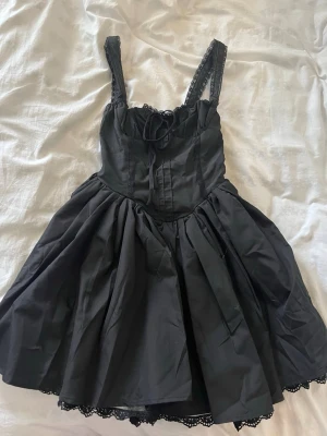 Svart kort klänning med spetsdetaljer - Supersöt svart kort klänning med spetsdetaljer på axelbanden och nederkanten. Klänningen har snörning framtill och en voluminös kjol som ger en riktigt fin siluett. Perfekt för dig som gillar en unik och lekfull stil.