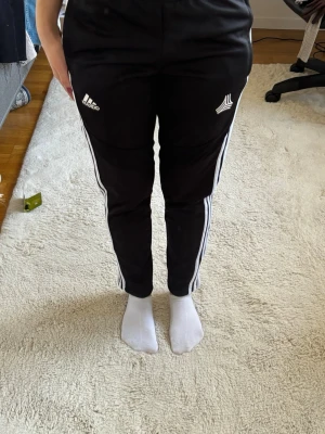 Svarta Adidas mjukisbyxor med vita ränder - Säljer ett par svarta mjukisbyxor från Adidas med klassiska vita ränder längs benen och logga på låret. Byxorna har smal passform, resår i midjan och är tillverkade i ett mjukt och stretchigt material som passar perfekt för chill eller träning. stadium 