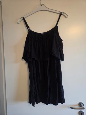 Svart volang topp med smala axelband, stl XL - Svart klänning med smala axelband och volangdetalj över bysten. Klänningen är ärmlös och har en luftig, loose passform som ger en avslappnad känsla. Perfekt för sommaren och enkel att styla med accessoarer.