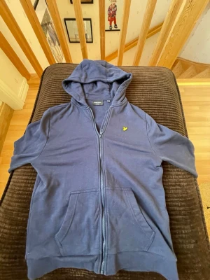 Blå hoodie från Lyle & Scott - Snygg blå hoodie från Lyle & Scott med dragkedja framtill och korta ärmar. Klassisk gul logga på bröstet och två stora fickor framtill. Perfekt för chill dagar och enkel att matcha med jeans eller shorts.ps knappt använd 