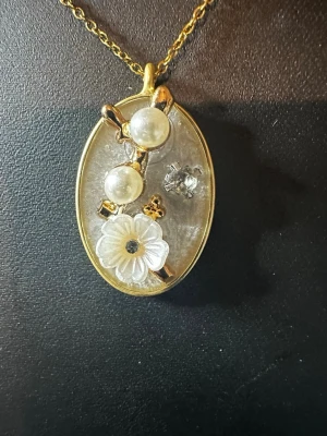 Guldigt halsband med pärlor och blomma - Unikt halsband med en oval guldtonad berlock, dekorerad med två vita pärlor, en genomskinlig blomma och små guldfärgade detaljer. Kedjan är tunn och elegant, perfekt för dig som gillar romantiska och kreativa smycken.