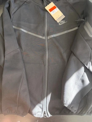 Nike Tech Fleece hoodie – svart (M/L) - Säljer en snygg svart Nike Tech Fleece hoodie. Den har dragkedja framtill och huva, och sitter riktigt bekvämt. De grå detaljerna över bröstet ger en stilren look, och det finns även en liten ficka med dragkedja på ärmen.  Passar perfekt till vardags eller om du vill ha en sportig stil.  Storlek: M–L  💬 Hör av dig om du är intresserad!