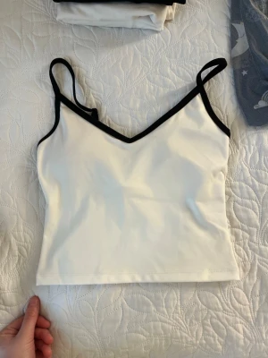 Vit linnetopp med svarta kanter - Snygg vit linnetopp med svarta kontrastkanter och smala axelband. Toppen har en v-ringad halsringning och croppad passform, perfekt till jeans eller kjol. Enkel och stilren design som passar till många olika looks.