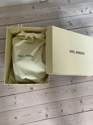 Svarta Axel Arigato sneakers - Snygga svarta sneakers från Axel Arigato i slät läder med minimalistisk design och vit sula. Skorna har diskret logga på sidan och kommer med originalförpackning och dustbag. Perfekta för dig som gillar stilrena och moderna sneakers.