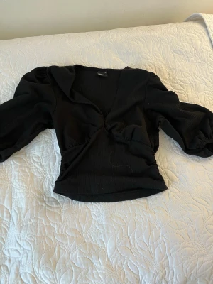 Svart v-ringad topp med puffärm - Trendig svart topp med v-ringning och puffiga långärmar. Modellen är croppad och har en åtsittande passform i midjan. Perfekt för dig som vill ha en stilren men ändå cool look.