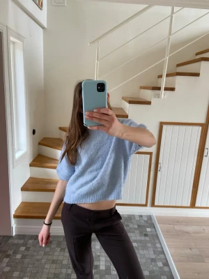 Ljusblå stickad kortärmad tröja - Mysig ljusblå stickad tröja med korta ärmar och ribbade kanter. Tröjan har en boxig, croppad passform och rund halsringning. Perfekt för lager på lager och enkel att styla till olika looks. Materialet ser ut att vara mjukt och fluffigt, troligen en ull- eller mohairblandning.