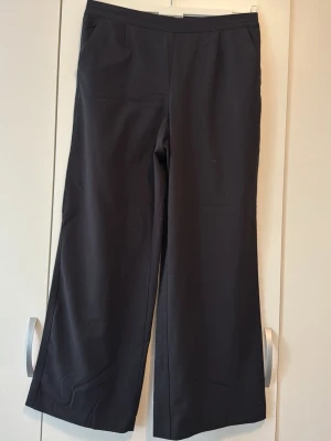 Svarta vida kostymbyxor - Snygga svarta kostymbyxor med hög midja och vida ben. Byxorna har resår i midjan baktill för extra komfort och två sidofickor framtill. Perfekta för en clean och stilren look. Materialet känns mjukt och följsamt.