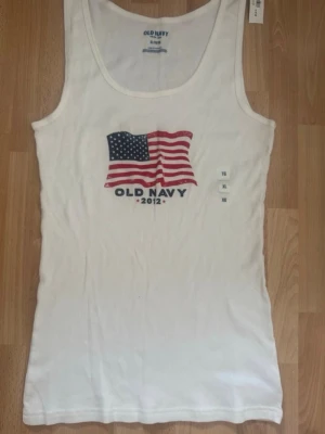 Ny vit ribbad linnetopp Old Navy - Säljer en vit ribbad linnetopp från Old Navy med tryck av amerikanska flaggan och texten 'Old Navy 2012' framtill. Toppen är ärmlös och har rund halsringning. Perfekt för sommardagar och enkel att matcha med jeans eller shorts. Köpt i USA, bomull 100%