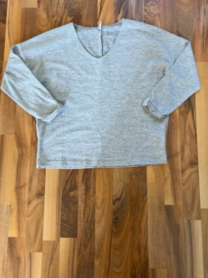 Grå V-ringad tröja från Hailys - Mjuk och enkel grå tröja från Hailys med V-ringning och långa ärmar. Tröjan har en avslappnad passform och är tillverkad i ett tunt, stretchigt material som känns skönt mot huden. Perfekt för lager på lager och en clean look.