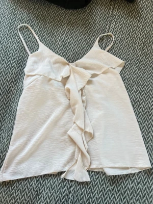 Vit volangtop med tunna axelband - Säljer en söt topp med tunna axelband och volangdetalj framtill. Toppen har en lätt v-ringad form och är tillverkad i ett mjukt, något glansigt tyg som känns luftigt. Perfekt för varma dagar och enkel att styla med jeans eller kjol.