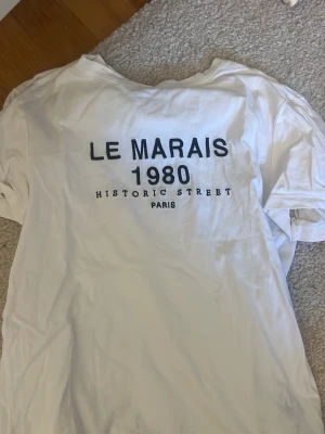 Zara tshirt  - Vit t-shirt med svart texttryck på ryggen där det står 'LE MARAIS 1980 HISTORIC STREET PARIS'. Klassisk passform och rund hals. Perfekt för dig som gillar enkel och stilren design med en touch av Paris.