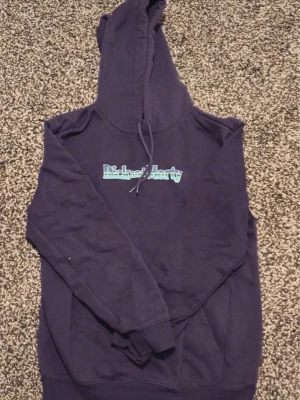 Mörklila Richard Harty hoodie - Säljer en mörklila hoodie från Richard Harty med tryckt logga i ljusblått och vitt på bröstet. Hoodien har klassisk huva med dragsko och ribbade muddar vid ärmslut och nederkant. Perfekt för dig som gillar streetwear och vill ha något bekvämt.