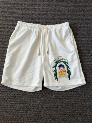 Casablanca shorts - Säljer ett par vita shorts från Casablanca med ett färgstarkt tryck framtill som föreställer en byggnad och gröna blad. Shortsen har elastisk midja med snörning och sidofickor. Perfekta för sommardagar och chill vibes.