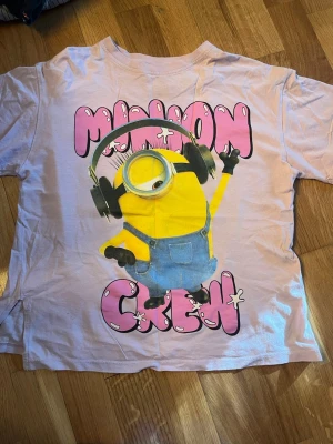 Ljusrosa/ljuslila Minions T-shirt - T-shirt i ljusrosa med stort Minions-tryck både fram och bak. Framsidan har texten 'Minion Crew' i rosa och en Minion med hörlurar, medan baksidan visar tre Minions i blå hängselbyxor. Perfekt för dig som älskar Minions och vill sticka ut.