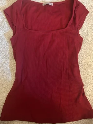 Röd ribbad topp med fyrkantig ringning - Snygg röd ribbad topp från Wild Field med fyrkantig ringning och korta ärmar. Toppen har en figurnära passform och är tillverkad i ett mjukt, stretchigt material som känns bekvämt mot huden. Perfekt att styla med jeans eller kjol.