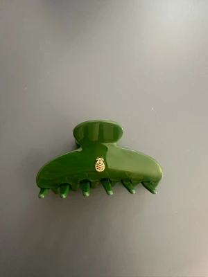 Grön hårklämma med ananasdetalj - Snygg grön hårklämma i plast med en liten guldfärgad ananasdetalj på ena sidan. Klämman har en klassisk form med bred öppning och flera piggar för att hålla håret på plats. Perfekt accessoar för att snabbt fixa frisyren.