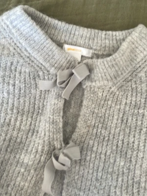 Ljusgrå stickad kofta med rosetter - Supersöt ljusgrå stickad kofta från Gina Tricot med fyra dekorativa rosetter framtill istället för knappar. Koftan har rund halsringning och är gjord i ett mjukt, fluffigt material som känns riktigt mysigt. Perfekt att slänga över en topp för extra stil.