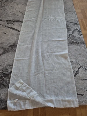 Vit gardinlängd i tunt tyg - Säljer en vit gardinlängd i tunt, lätt genomskinligt tyg med diskreta invävda ränder. Gardinen har kanal upptill för enkel upphängning på stång. Passar perfekt för att släppa in ljus men ändå ge lite insynsskydd.