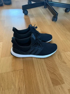 Svarta Adidas Ultraboost sneakers - Säljer ett par svarta Adidas Ultraboost sneakers med stickad ovandel och vita Boost-sulor. Skorna har klassisk låg profil, svarta snören och diskreta Adidas-ränder på sidan. Perfekta för dig som gillar stilrena och bekväma sneakers.
