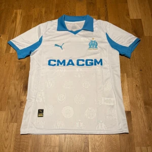 Marseille home kit 25/26 - Snygg vit matchtröja från Olympique de Marseille med blå detaljer och logga på bröstet. Tillverkad av Puma med CMA CGM sponsortryck på framsidan. Klassisk krage och korta ärmar, perfekt för fotboll eller träning.