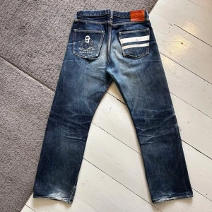 Mörkblåa Momotaro Selvedge Jeans - Snygga Momotaro jeans med battle stripes samt en väldigt sällsynt print på Momotaro-figuren på bakfickorna. | W31 ≈L32 | Nypris: ≈3500kr | Bra skick förutom några uppsläppta sömmar på ena bakfickan, lite heelbite samt lite slitage i skrevet. Funkar utmärkt att bära dem som dem är, men annars kan man billigt fixa det hos skräddare. | Ytterlängd: 100cm, midja: 40cm, fotvidd: 21cm, innerbenslängd: 73cm.