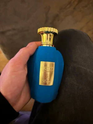 Voix The Duke parfym från Emor - Lyxig parfymflaska från Emor, modellen Voix The Duke. Flaskan är klädd i sammetsliknande blått material med guldfärgad metallkork och guldfärgad etikett framtill. Elegant och unik design som sticker ut i parfymhyllan.