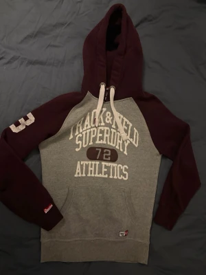 Superdry Track & Field hoodie - Snygg hoodie från Superdry med grå kropp och vinröda ärmar och huva. Stor tryckt text på bröstet med 'Track & Field Superdry Athletics 72'. Huva med vita dragsnören och känguruficka framtill. Perfekt för en sportig och avslappnad stil.🚨priset kan alltid diskuteras🚨skriv om ni har frågor!