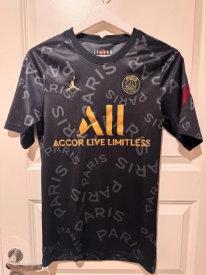 Svart PSG x Jordan matchtröja - Snygg svart PSG matchtröja i samarbete med Jordan. Tröjan har guldiga detaljer, PSG-logga och Jordan-logga på bröstet samt texten 'PARIS' tryckt över hela. Tillverkad i lätt och ventilerande material, perfekt för träning eller match.