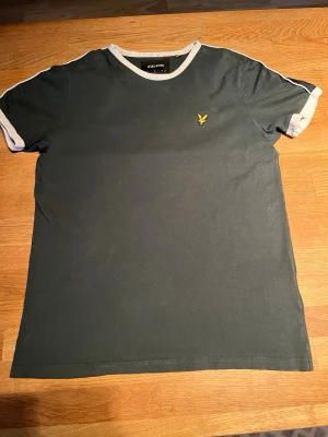 Grön t-shirt från Lyle & Scott - Mörkgrön t-shirt från Lyle & Scott med vit kant vid hals och ärmslut. Klassisk gul logga på bröstet. Skön passform och tillverkad i mjuk bomull, perfekt till jeans eller shorts.