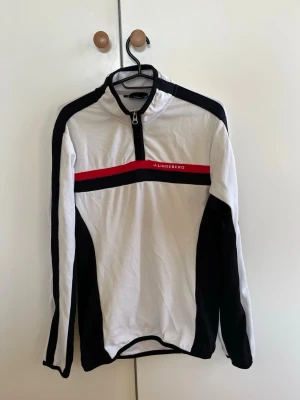 Vit och svart sporttröja J.Lindeberg - Sportig långärmad tröja från J.Lindeberg i vitt och svart med en röd detalj och logotyp på bröstet. Tröjan har halv dragkedja framtill och är tillverkad i ett lätt och funktionellt material som passar perfekt för träning och aktivitet eller till vardags. Nypris 1500 kr. Bra skick och storlek L men sitter som en Medium