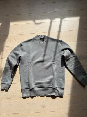 Grå sweatshirt med rund hals - En stilren grå sweatshirt med rund hals och ribbade muddar vid ärmslut och nederkant. Tröjan har en enkel och avslappnad passform, perfekt för en casual look. Tillverkad i mjukt material som känns skönt mot huden.