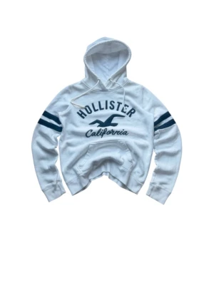Vit Hollister hoodie med blå detaljer XS - Snygg vit hoodie från Hollister med blå tryck på bröstet och två blå ränder på varje ärm. Klassisk huva med dragsko och magficka framtill. Mjuk och skön bomullsblandning, perfekt för en avslappnad stil.