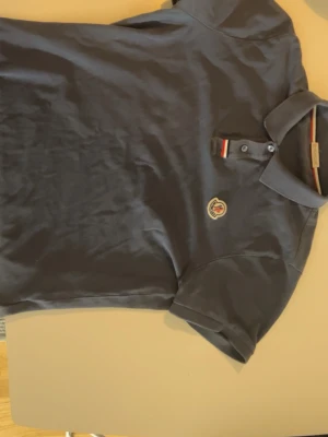 Mörkblå Moncler pikétröja - Snygg mörkblå pikétröja från Moncler med klassisk krage, korta ärmar och knappslå med rödvit detalj. Tröjan har Monclers ikoniska logga på bröstet och är tillverkad i mjuk bomull. Perfekt till jeans eller shorts för en clean look.