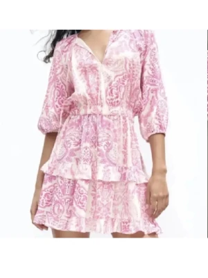 Rosa och vit kortklänning med volang - Supersöt kortklänning i rosa och vitt med blommigt mönster och volangdetaljer. Klänningen har v-ringning, trekvartsärm och resår i midjan för en bekväm passform. Perfekt för sommarens alla tillfällen!