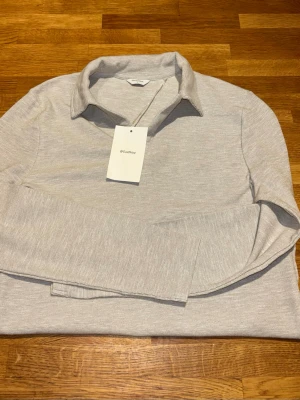 Ljusgrå långärmad pikétröja - Ljusgrå långärmad pikétröja från EastWest med klassisk krage och diskret struktur i tyget. Tröjan har en clean och stilren look, perfekt för dig som gillar minimalistisk stil. Tillverkad i mjukt material som känns bekvämt hela dagen.