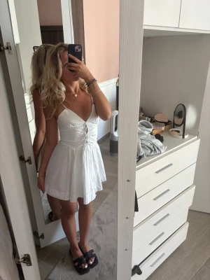 Vit kort sommarklänning med snörning - Söt vit kortklänning med smala axelband och snörning framtill. Klänningen har v-ringning och en lätt utställd kjol som ger en luftig känsla. Perfekt för varma dagar och har en enkel, fräsch look.