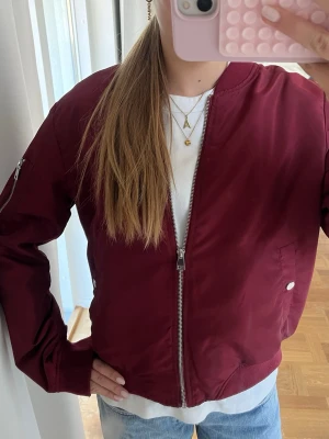 Vinröd bomberjacka från Vero Moda - Snygg vinröd bomberjacka från Gina Tricot med silvriga dragkedjor och tryckknappar. Jackan har ribbade muddar vid ärmslut och nederkant samt fickor på sidorna och en extra ficka på ärmen. Tillverkad i glansigt material som ger en cool look.