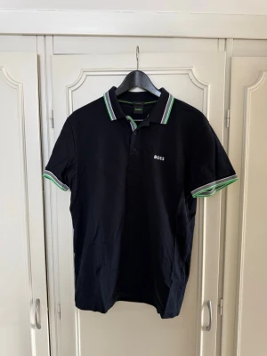 Svart kortärmad piké från BOSS - Snygg svart pikétröja från BOSS med vita och gröna detaljer på krage och ärmslut. Klassisk passform med korta ärmar och liten logga på bröstet. Tillverkad i mjuk bomull som känns skön mot huden. Perfekt för en clean och stilren look.