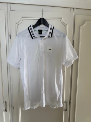 Vit kortärmad t-shirt från BOSS - Vit kortärmad t-shirt från BOSS med svartvitrandig krage och liten BOSS-logga på bröstet. Klassisk pikédesign med knappar vid halsen. Tillverkad i mjuk bomull för en bekväm känsla och stilren look.