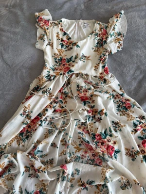 Vit blommig långklänning med volang - Supersöt långklänning i vit bas med blommigt mönster i rosa, grönt och gult. Klänningen har korta volangärmar, v-ringning och knytband i midjan för en feminin siluett. Tillverkad i ett lätt och luftigt tyg, perfekt för varma dagar.