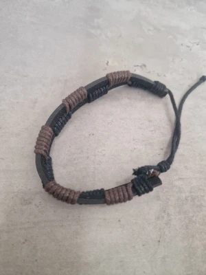 Svart och brunt justerbart armband - Snyggt armband i svart och brunt med justerbar knytning. Armbandet är gjort av svart läderimitation och dekorerat med bruna och svarta tvinnade trådar i sektioner runt hela bandet. Perfekt accessoar för en avslappnad och trendig stil.