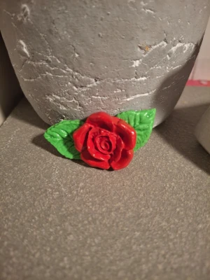Röd rosformad brosch i lera - Säljer en unik handgjord brosch i form av en röd ros med två gröna blad. Broschen är tillverkad i lera och har en blank finish som ger ett snyggt intryck. Perfekt för att piffa upp jackan eller väskan med en färgklick och lite romantisk vibe.