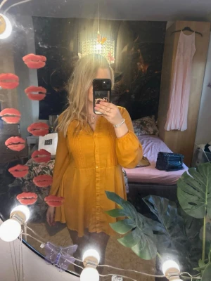 Gul kortklänning med knappar - Säljer en söt gul kortklänning med knappar framtill och långa ärmar. Klänningen har en lätt v-ringning och fina veckdetaljer på bröstet. Perfekt för dig som gillar färg och vill ha en bekväm men snygg look.