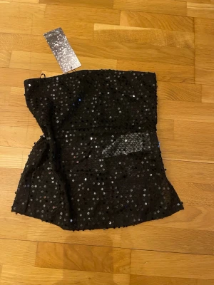 Svart paljett-tubtopp - Snygg svart tubtopp stl.s täckt med glittrande paljetter. Toppen är axelbandslös och har en figurnära passform över brösten och lösare nertill. Perfekt för dig som vill ha något extra till din outfit.