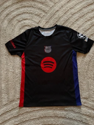 Svart FC Barcelona matchtröja - Snygg svart matchtröja från FC Barcelona med röda och blå detaljer på sidorna. Tryck med Spotify-logga på bröstet och Lamine Yamal samt nummer 10 på ryggen. Korta ärmar och klubbmärke på bröstet. Perfekt för dig som älskar fotboll och Barca.