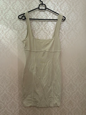 Beige kort klänning med breda axelband - Stilren beige kort klänning med breda axelband och fyrkantig urringning. Klänningen har markerade sömmar framtill som ger en snygg siluett. Perfekt för dig som gillar minimalistisk och clean stil.
